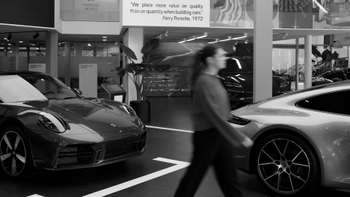 Interior de un showroom Porsche con dos modelos 911, una persona en movimiento y una cita de Ferry Porsche en la pared.