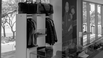 Exposición de ropa y accesorios Porsche en ventanal de showroom, con bolsas y perchas de prendas, y una gráfica de personas en la pared.