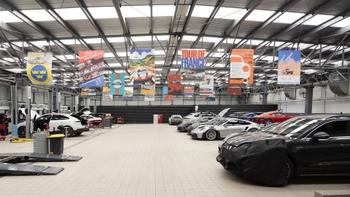 Amplio taller de servicio Porsche con múltiples vehículos, elevadores y banners conmemorativos de carreras y eventos colgando del techo.