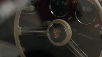 Primer plano del volante beige de un coche clásico Porsche, mostrando el escudo de la marca y parte del salpicadero con indicadores.