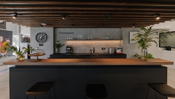 Área de cocina y café "Porscheplatz" en el concesionario Porsche Centre Santa Fe, con barra de madera, cafetera, utensilios y plantas ornamentales.