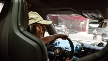 Mujer sonriendo al volante de un Porsche, vista desde el asiento trasero mostrando el salpicadero y el logo en el volante.