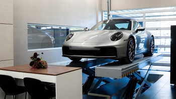 Un Porsche 911 plateado exhibido sobre un elevador en el interior de un moderno concesionario en Tijuana, con una mesa y arreglo floral.