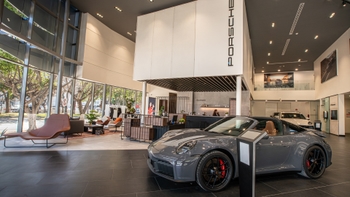 Un Porsche 911 gris en el primer plano del moderno interior de un concesionario Porsche, con recepción y lounge a través de amplias ventanas.