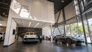 Amplio interior del concesionario Porsche Centre Tijuana con un Macan blanco y un 911 descapotable gris. Una cita de Ferry Porsche en inglés decora la pared.