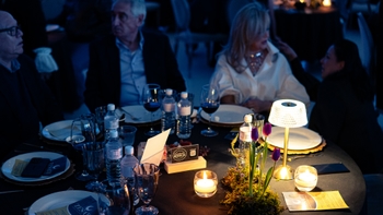 Mesa iluminada en cena de gala con velas, florero y copas; invitados conversando en segundo plano.