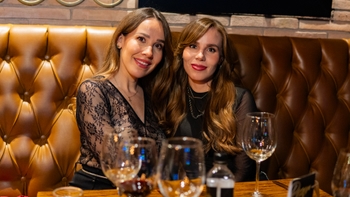 Dos mujeres sonrientes posando en una mesa durante una cena, con copas y ambiente cálido.