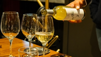 Primer plano de vino blanco siendo servido en una copa, con otras copas y cristalería en la mesa.