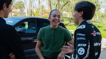Mujer sonriente conversando con un piloto de carreras junto a un Porsche oscuro en un entorno exterior.