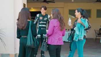 Joven piloto de carreras con uniforme de Porsche rodeado de mujeres conversando al aire libre.