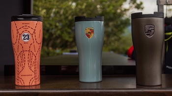 Tres vasos térmicos alineados sobre una mesa, con el emblema Porsche en el vaso central.