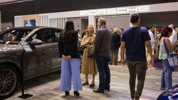 Asistentes interactuando y conversando en un evento de Porsche, con un SUV Porsche visible y el logo de la marca al fondo.