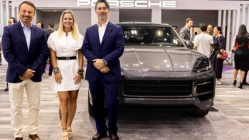 Tres personas posando junto a un SUV Porsche gris en la presentación de un evento, con el logo "PORSCHE" visible al fondo.