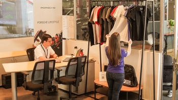 Mujer mirando ropa Porsche Lifestyle en una tienda, con un empleado trabajando en un escritorio en el fondo.