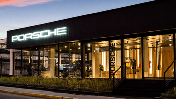 Vista nocturna de la fachada de un concesionario Porsche con grandes ventanales y el logo iluminado en la parte superior.