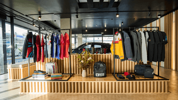 Interior de tienda de concesionario Porsche con exhibición de ropa, accesorios y un auto visible al fondo.