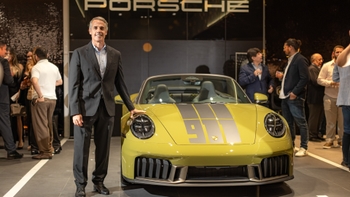 Porsche 911 descapotable amarillo con franjas y número en el capó, expuesto en un showroom; una persona de traje posa junto al coche y hay asistentes al fondo