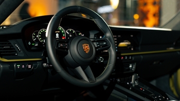 Interior del Porsche 911 en primer plano: volante con el escudo Porsche, indicadores digitales y controles del tablero visibles.