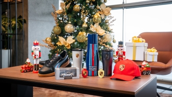 Variedad de productos de merchandising de Porsche presentados sobre una mesa con un árbol de Navidad al fondo, incluyendo zapatos, gorras, perfumes y modelos a escala.
