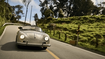 Un clásico Porsche 356 Speedster de color crema o beige claro se desplaza por una carretera de montaña sinuosa con una línea amarilla central. El coche convertible tiene los faros encendidos y está rodeado por un paisaje verde exuberante y arbolado bajo un cielo parcialmente nublado.